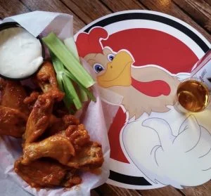 Sauced chicken wings with celery and dipping sauce beside the Rooster's logo.