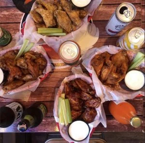 Overhead view of four chicken wing baskets with celery, dipping sauces, beer, hard seltzer and bottled soda.