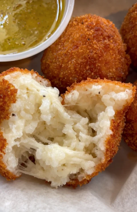 Homemade rice balls, one cut open to show rice and melted cheese, with dipping sauce.