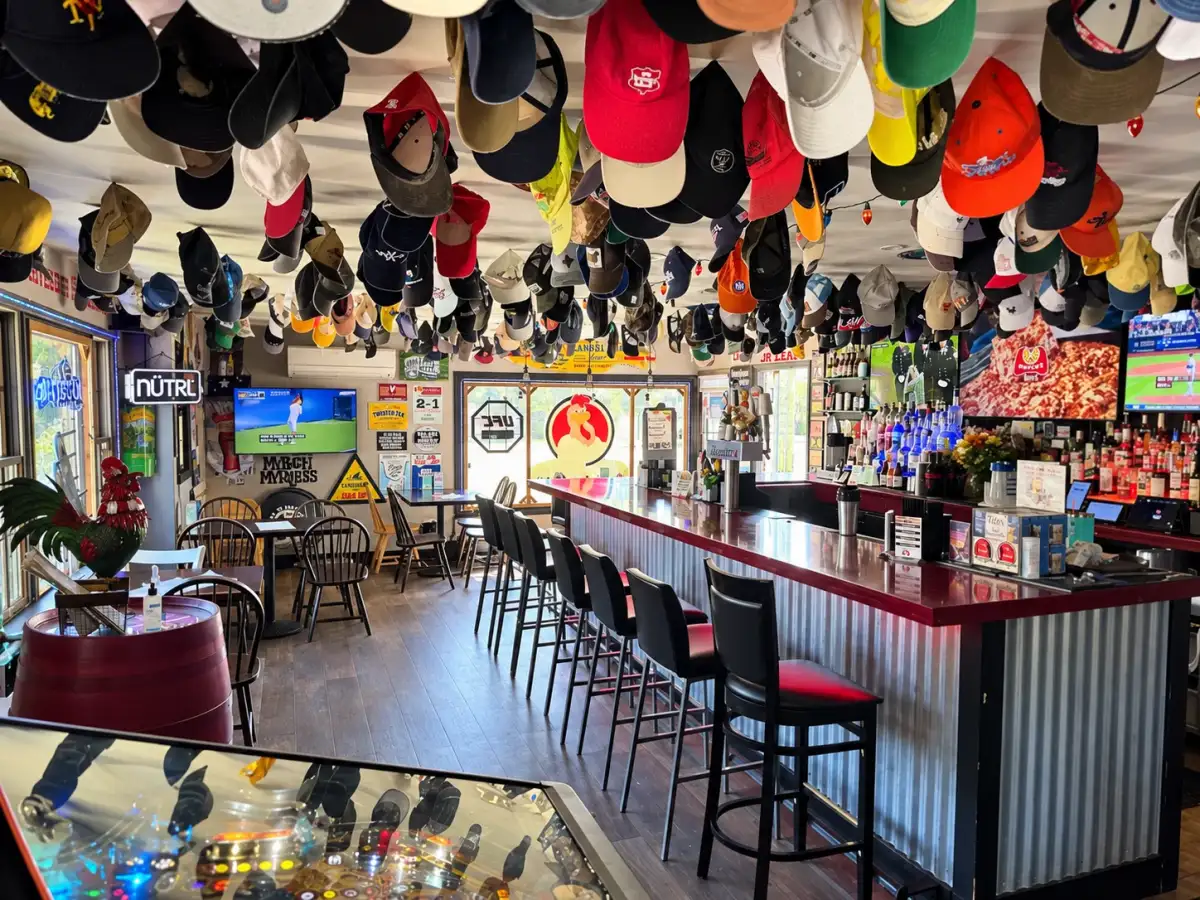 The full bar at Rooster's Wings & Brew, with bar seating, TVs and baseball caps hanging from the ceiling.